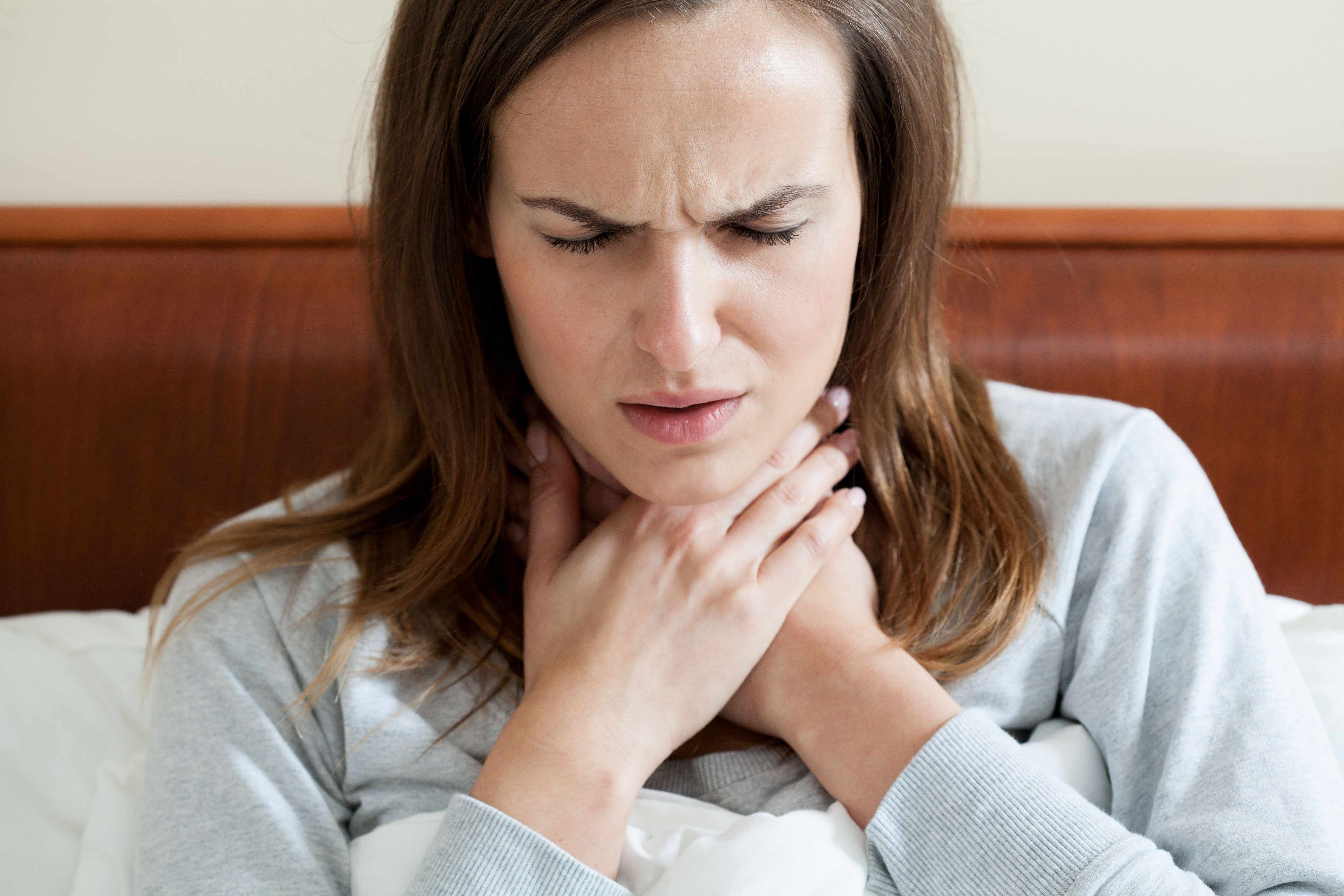 a Caucasian woman sitting in bed holding her neck in pain with a sore throat