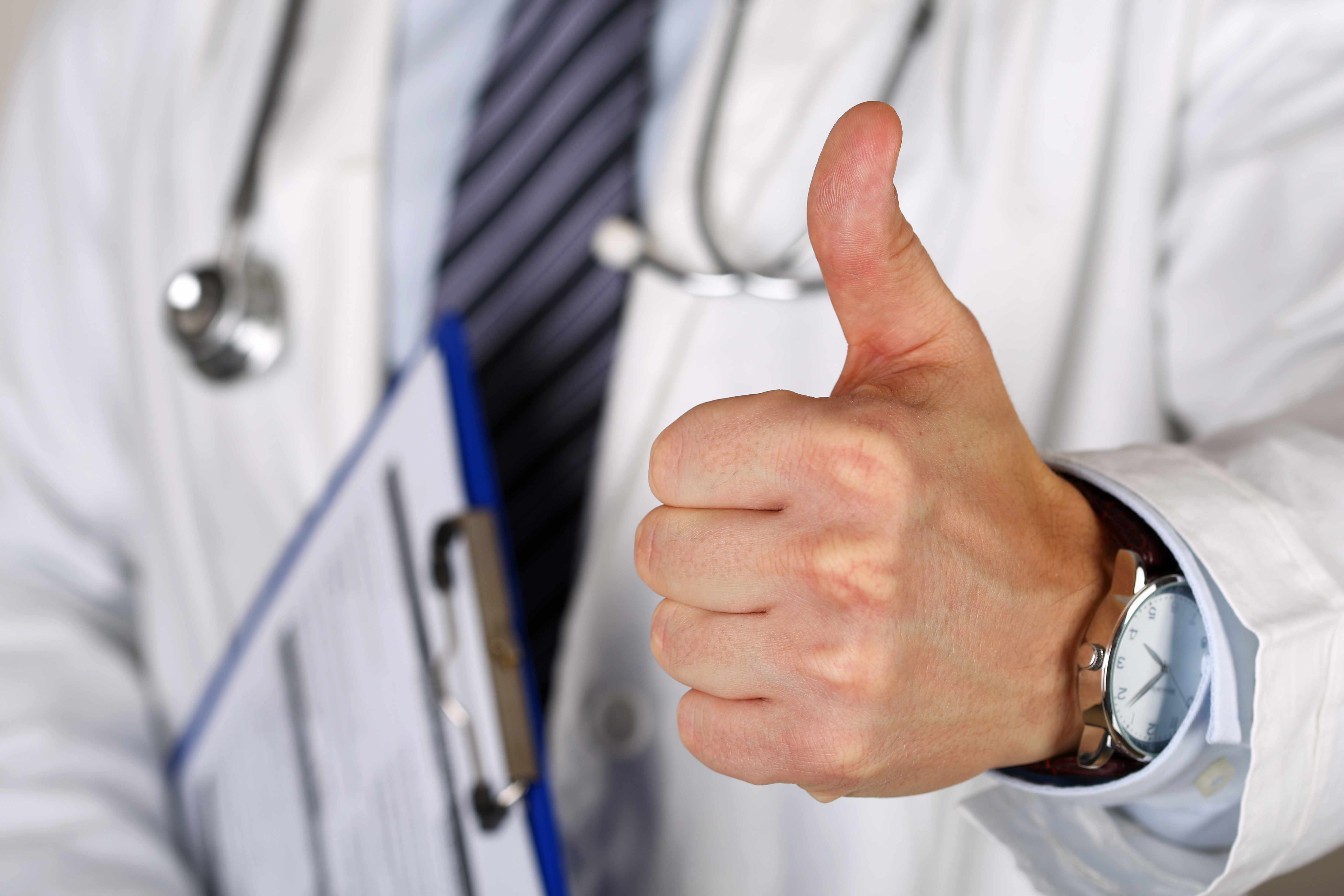 a medical staff person in a white coat and wearing a stethoscope giving the approval thumbs up sign