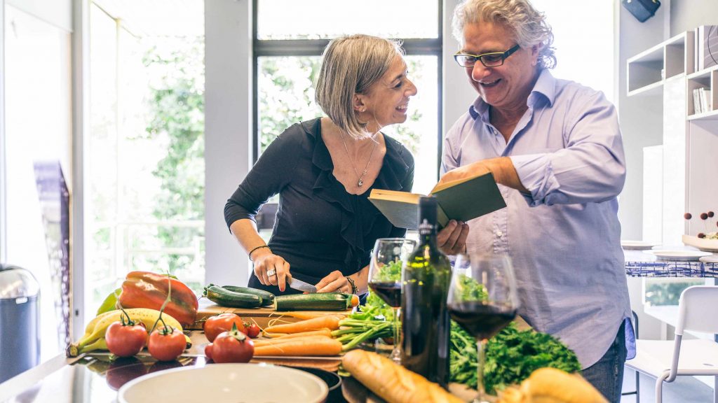 Una pareja de mediana edad sonríe y cocina en conjunto, mientras hay muchas frutas y verduras diseminadas sobre la barra de la cocina