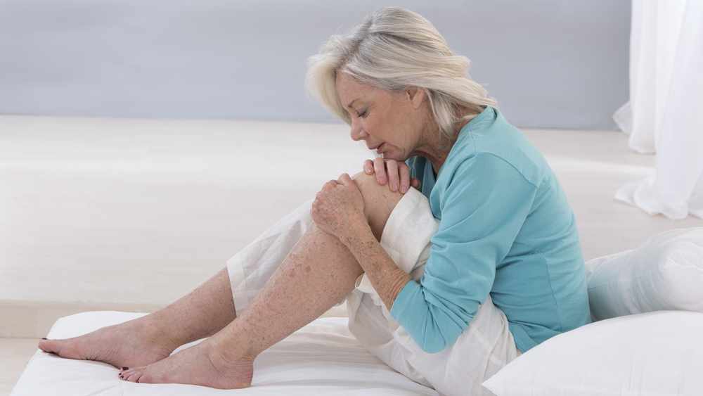 una anciana sentada en la cama hace muecas de dolor mientras se sostiene la rodilla