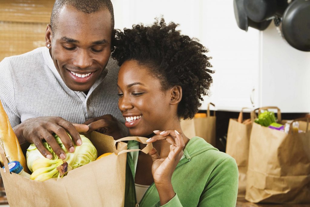 a smiling young couple in their kitchen, looking into a bag of groceries