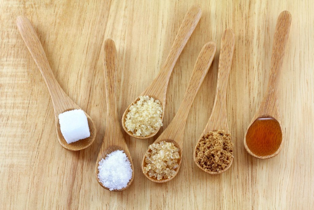 several wooden spoons full of different types of sugar, on a wooden tabletop