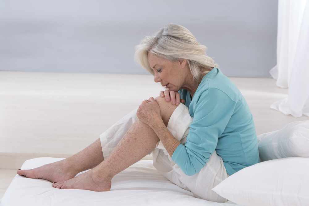 an older woman sitting on a bed, grimacing in pain and holding her knee