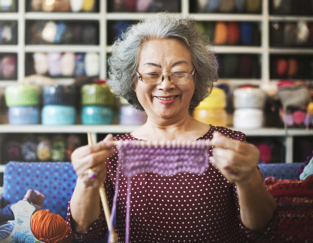 a smiling older woman holding up a knitting project, against a wall of yarn and craft supplies