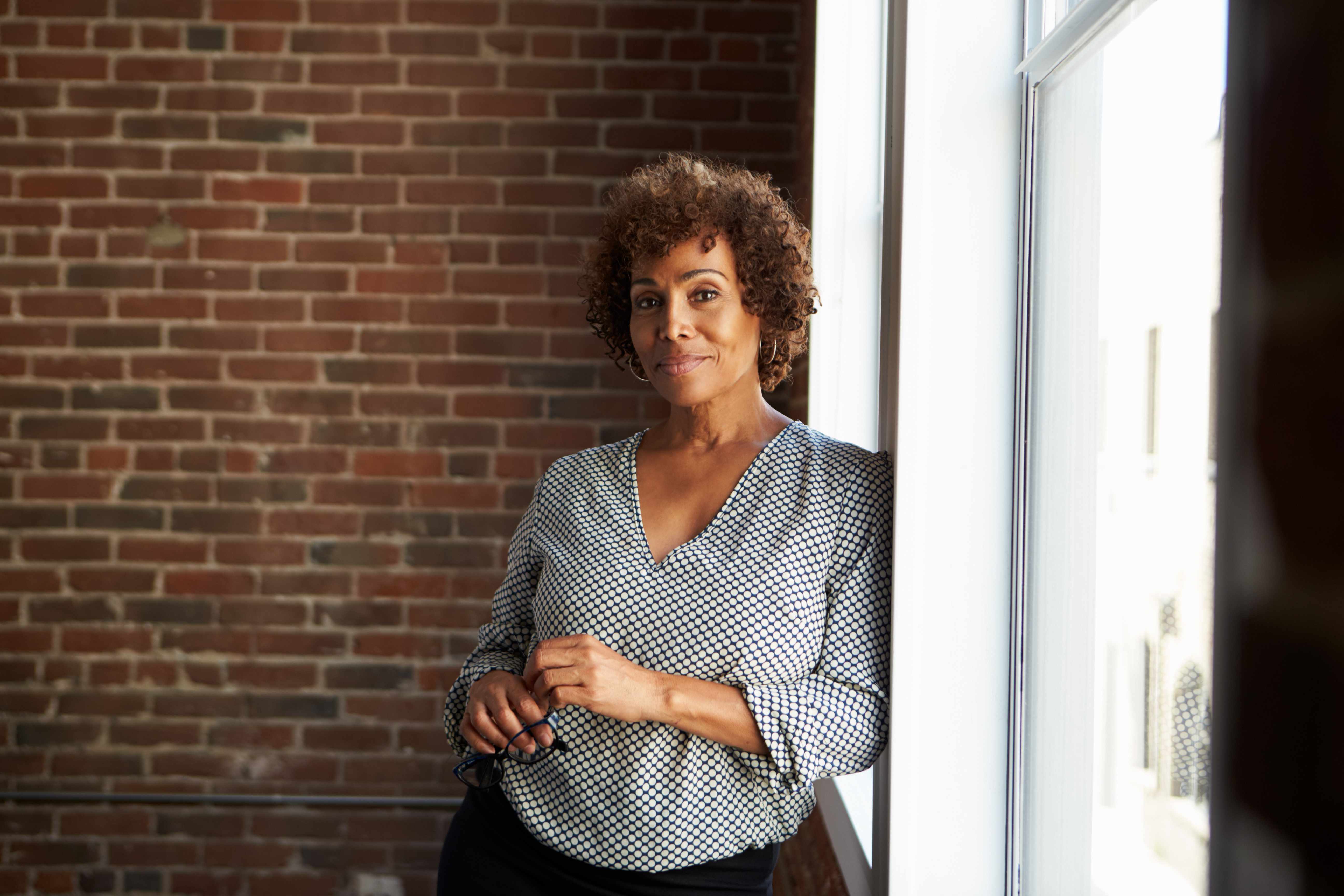 a middled aged African-American woman in casual business outfit, leaning against a window with brick wall in background