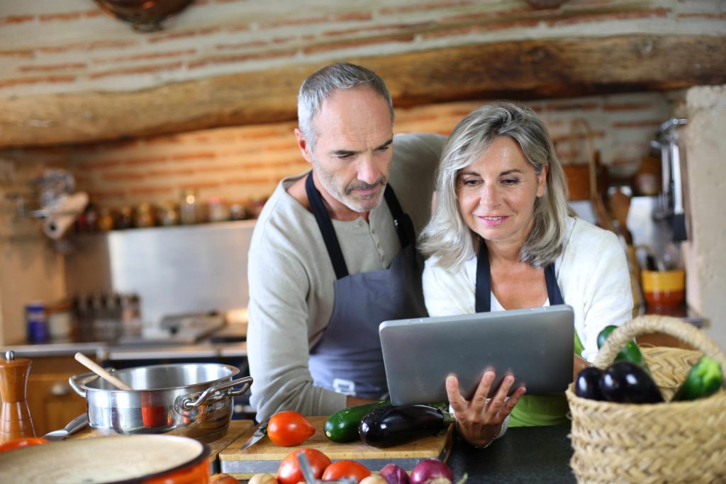 a middle-aged couple in a kitchen, looking at a laptop and preparing food