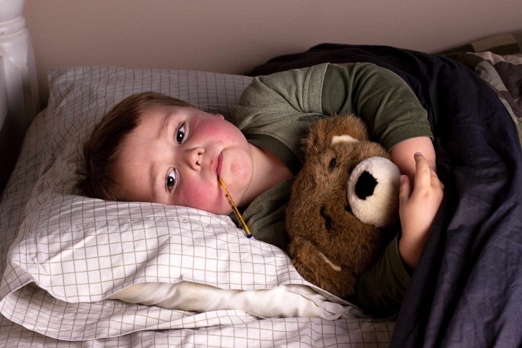 a sick little boy lying in bed, with red cheeks and a thermometer in his mouth, hugging a teddy bear