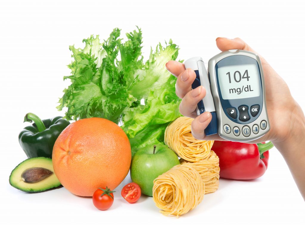 a hand holding a glucose meter in front of a variety of fruits, vegetables and pasta