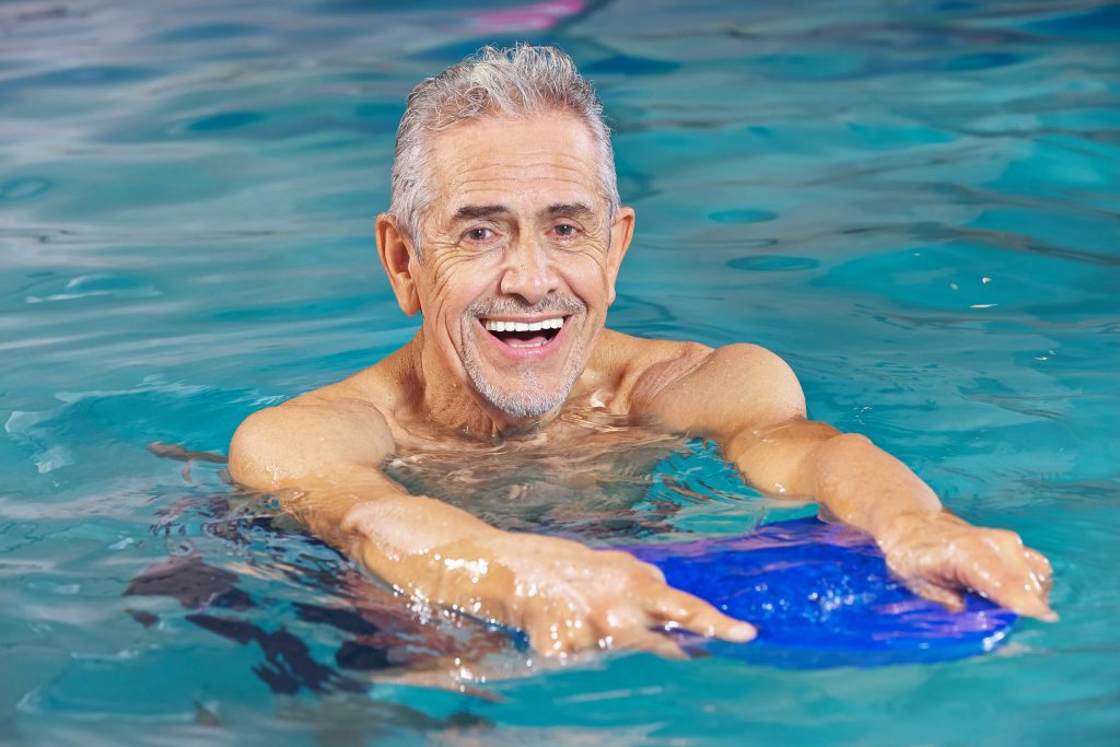 a close-up of a smiling older man in a swimming pool, holding a kickboard