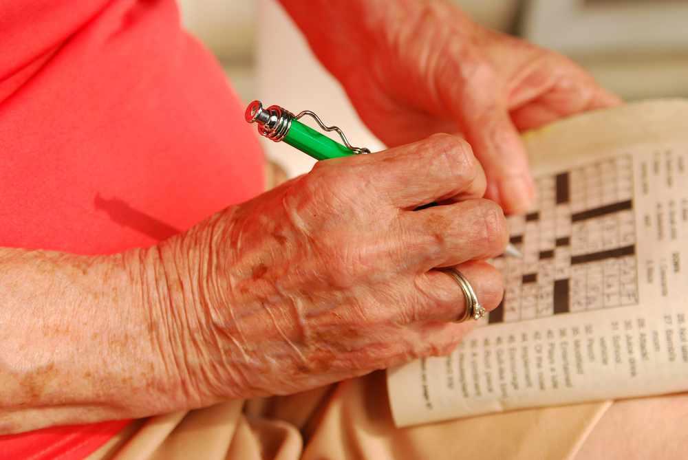 a close-up of an older woman's hand, holding a pen and working on a crossword puzzle