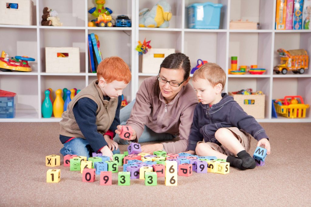 a mother and two young boys playing with blocks in a playroom, with orderly shelves of toys in the background