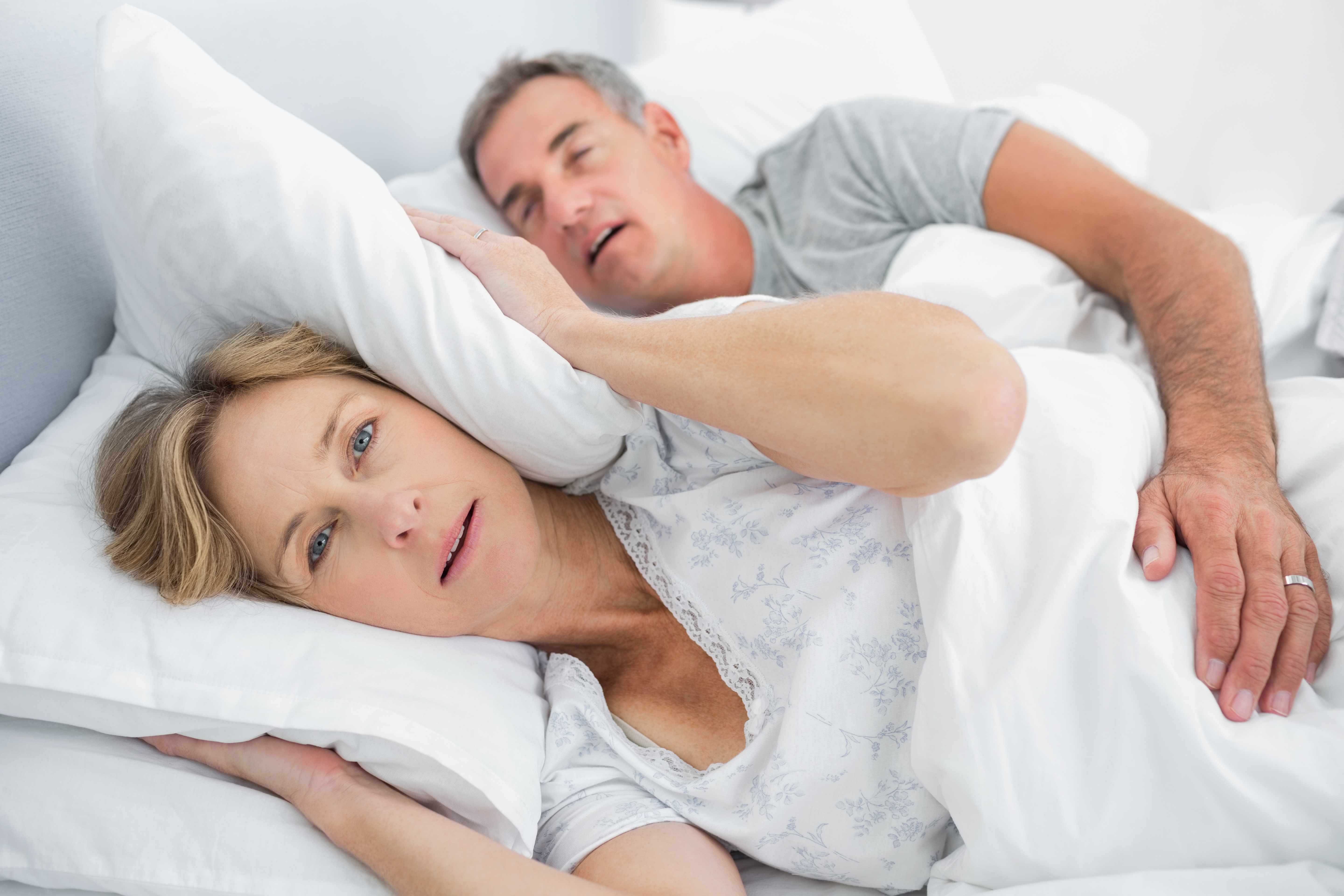 a middle-aged couple in bed with man snoring and woman covering her ears with a pillow