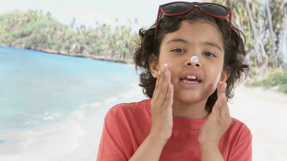 Aumenta el cáncer de piel: un niño muy simpático se aplica protector solar en la cara mientras está en la playa
