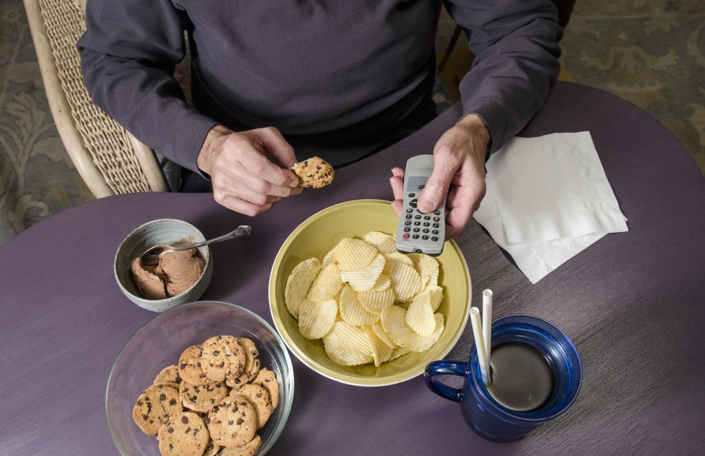 man sitting at table overeating, eating junk food, poor nutrition, binge eating, remote, TV