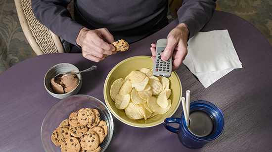 Un hombre sentado a la mesa come en exceso comida chatarra, que es mala nutrición, y se da un atracón, mientras sostiene el control remoto del televisor. 