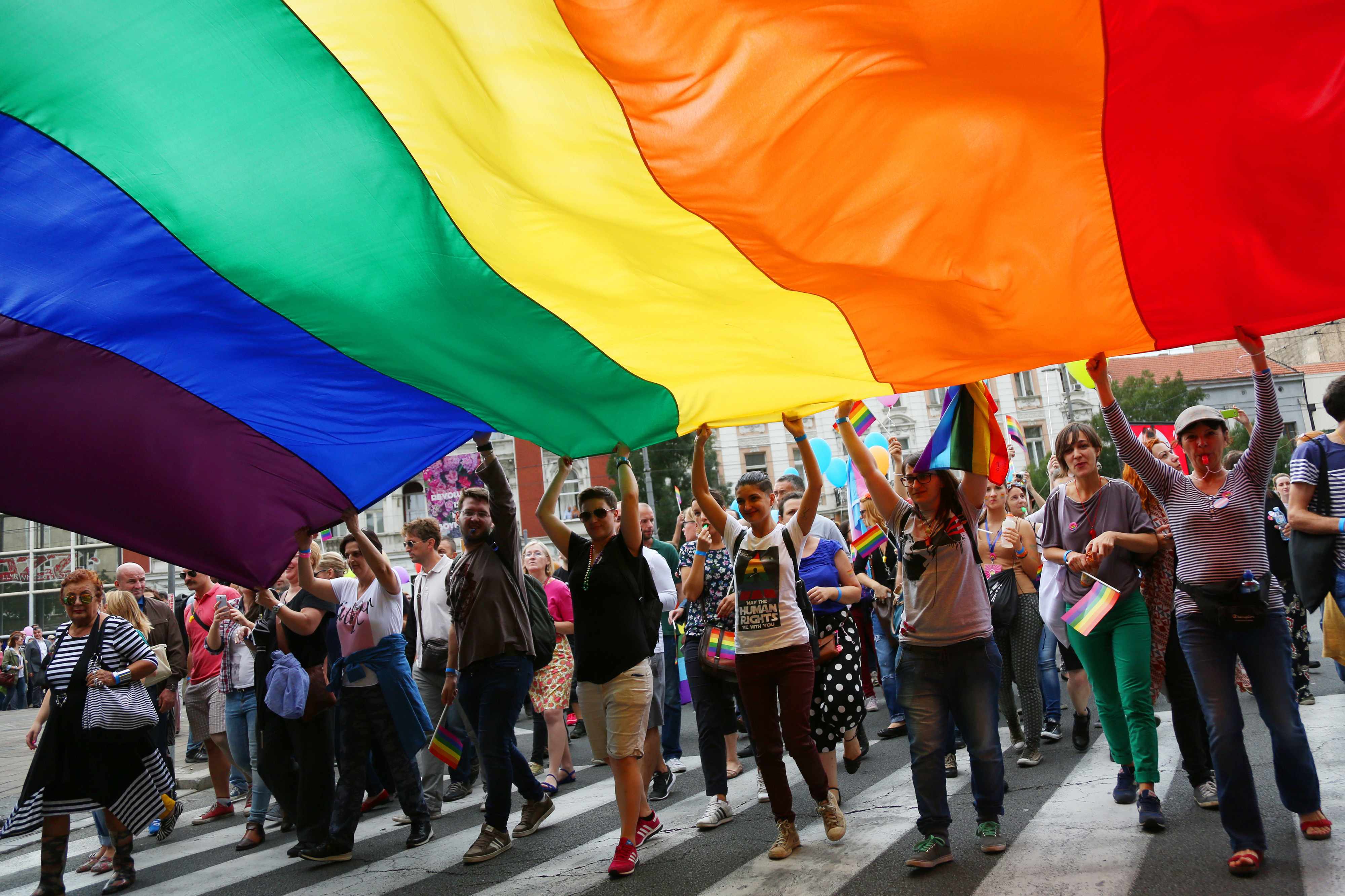 a group of smiling people in a Gay Pride parade, holding a large rainbow banner