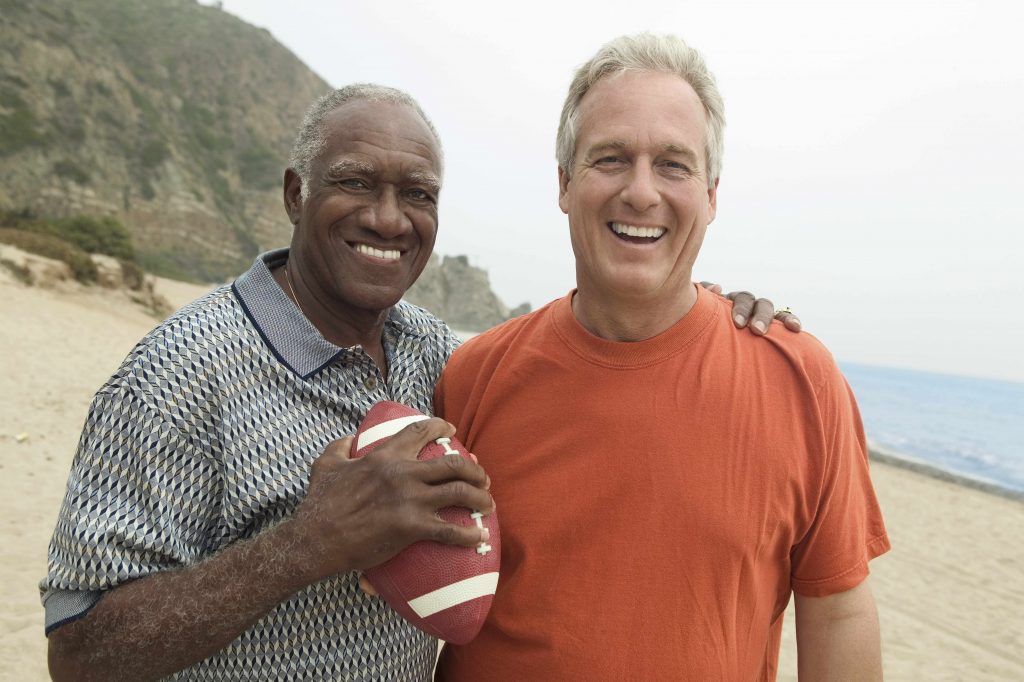 two smiling men standing together on a beach, one holding a football in one hand with the other arm around his friend's shoulders