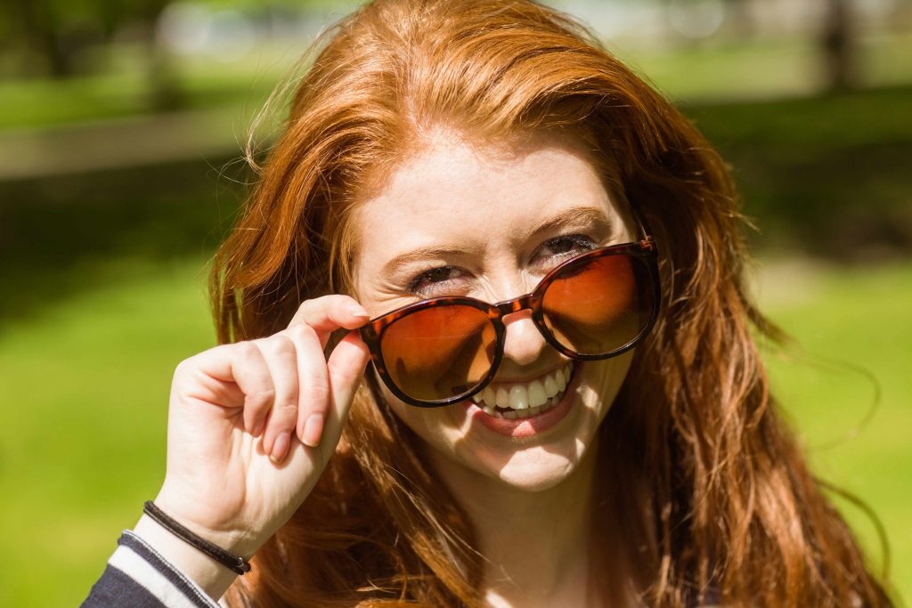 a smiling woman outside on a sunny day, peering over her sunglasses