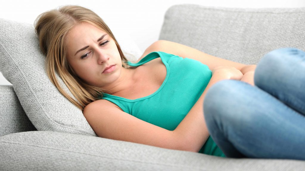 a young, sad looking woman lying on a couch holding her stomach as if she has a tummy ache, maybe cramps from her monthly period, menstrual cycle