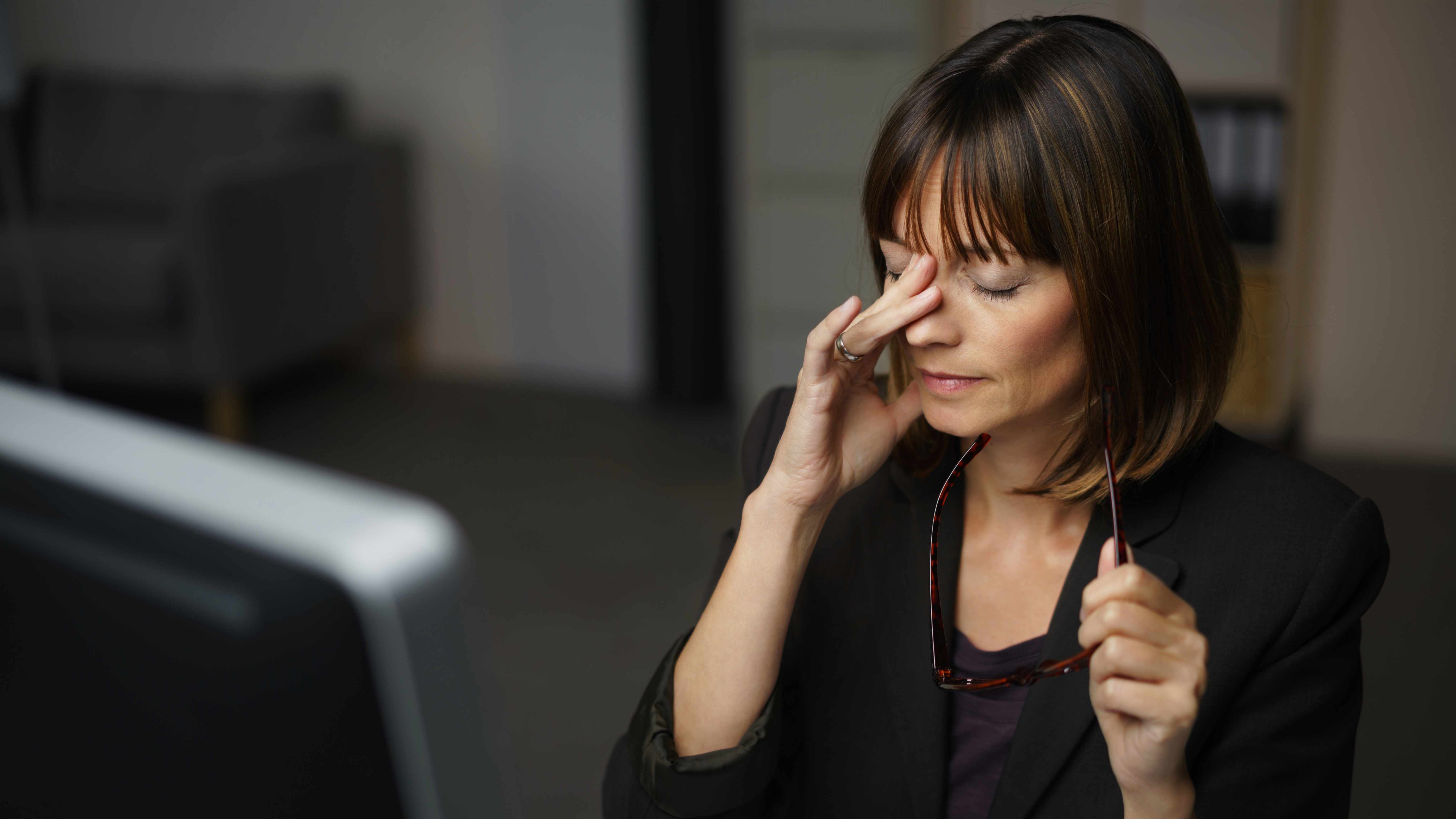 a young woman sitting at a computer in an office looking tired and touching her face like she has a headache
