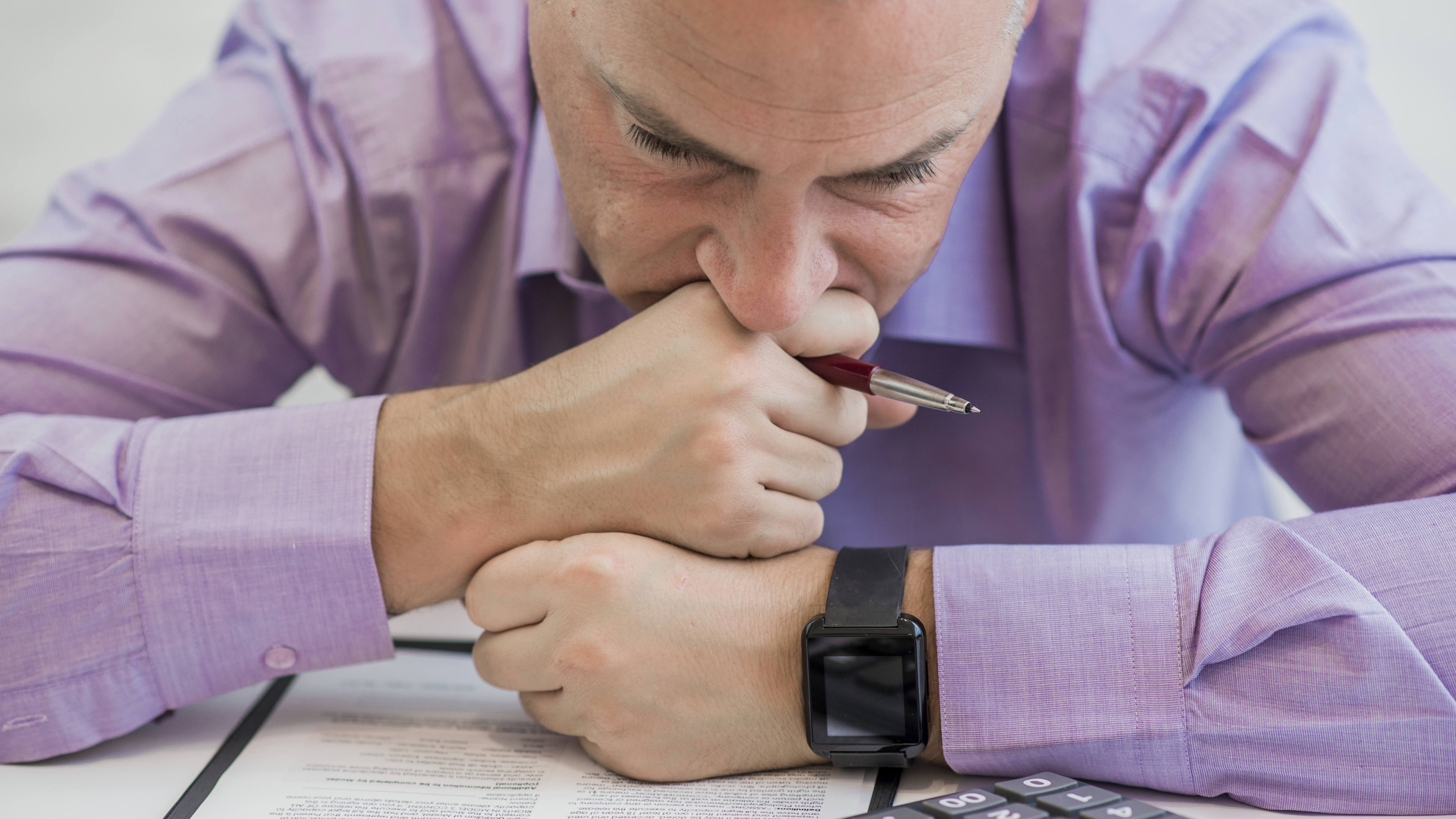 a man resting his head on his hands, holding a pen at work and looking sad, depressed or stressed in the office
