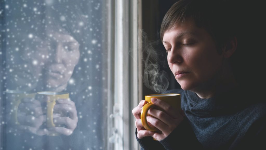 a middle-aged woman looking sad, with her eyes closed, holding a cup in both hands, standing in front of a dark window with snow falling outside