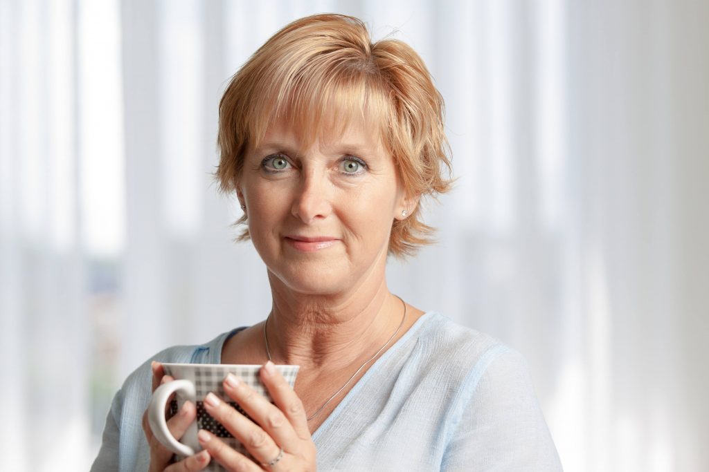 a close-up of an older woman, holding a cup in her hands, looking concerned, smiling tentatively and looking straight into the camera