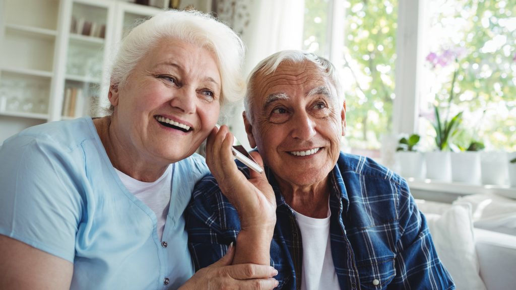 an older man and woman smiling and listening together to a cell phone