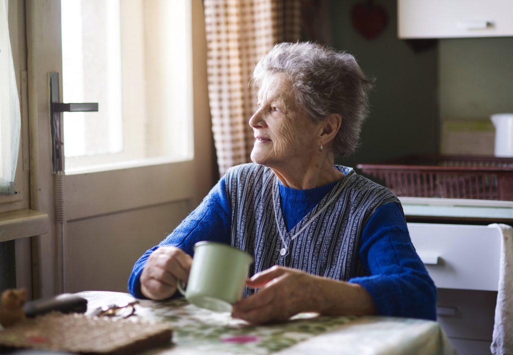 an elderly woman sitting at her kitchen table with a cup in hand, staring out the window