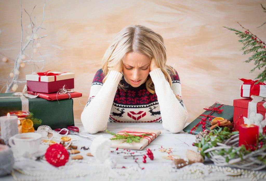 a woman tired and stressed during the holidays, holding her head in her hands with presents and wrapping paper everywhere