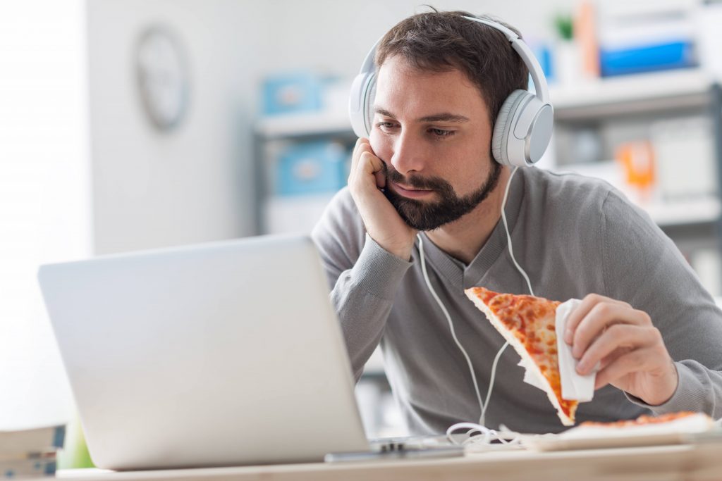 a young man alone in his work office, eating a slice of pizza and staring at his computer screen