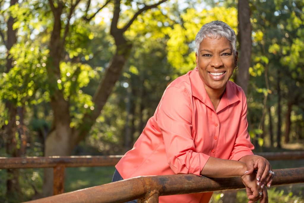 a happy, smiling middle-aged woman outside in a wooded area, leaning on a railing