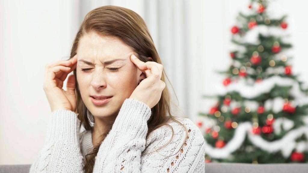 with a tree in the background, a woman holding her hands to her head as if she's tired or stressed, with a headache at the Christmas holidays