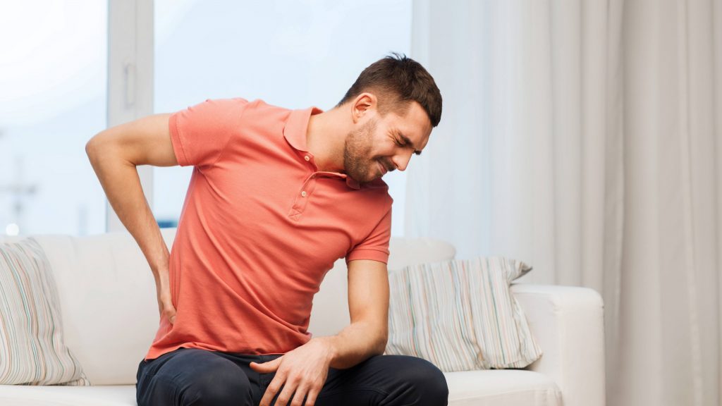 Un hombre sentado en un sofá se lleva la mano a la espalda en una muestra de dolor