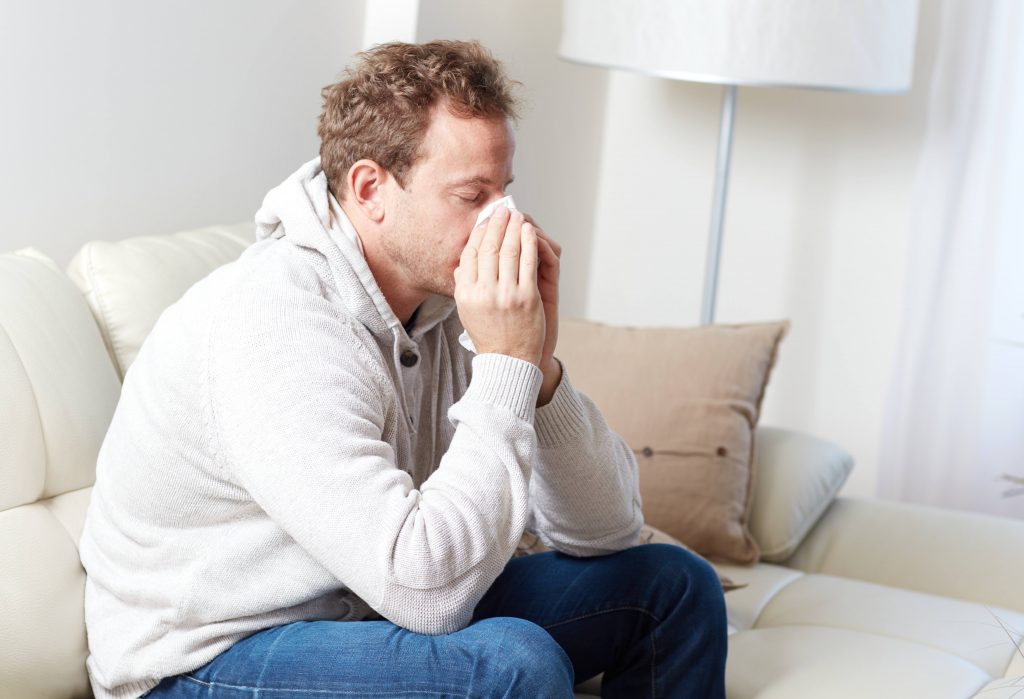 a young man sitting on a couch blowing his nose, with a cold, sinus allergies or a flu bug