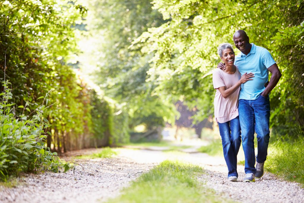 a happy, smiling middle-aged couple walking with their arms around one another down a dirt path, surrounded by trees and sunshine