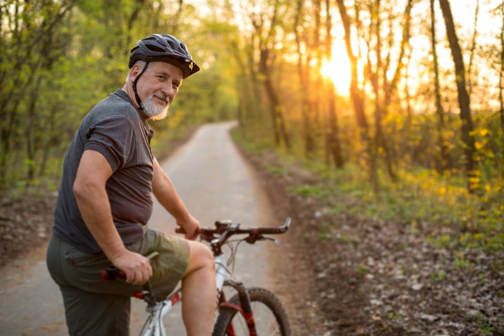 a smiling middle-aged man on a bike on a wooded bike path with the sun shining through the trees