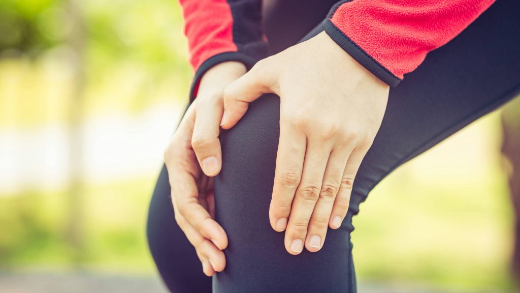 Una mujer en traje de ejercicio que sostiene el área de la pierna y la rodilla como si tuviera dolor después de una lesión.
