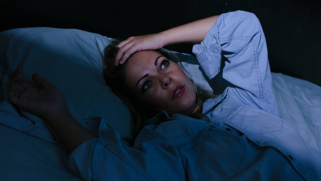 Una mujer joven está acostada en una habitación oscura, con los ojos abiertos y las manos sobre la frente porque sufre de insomnio y estrés.