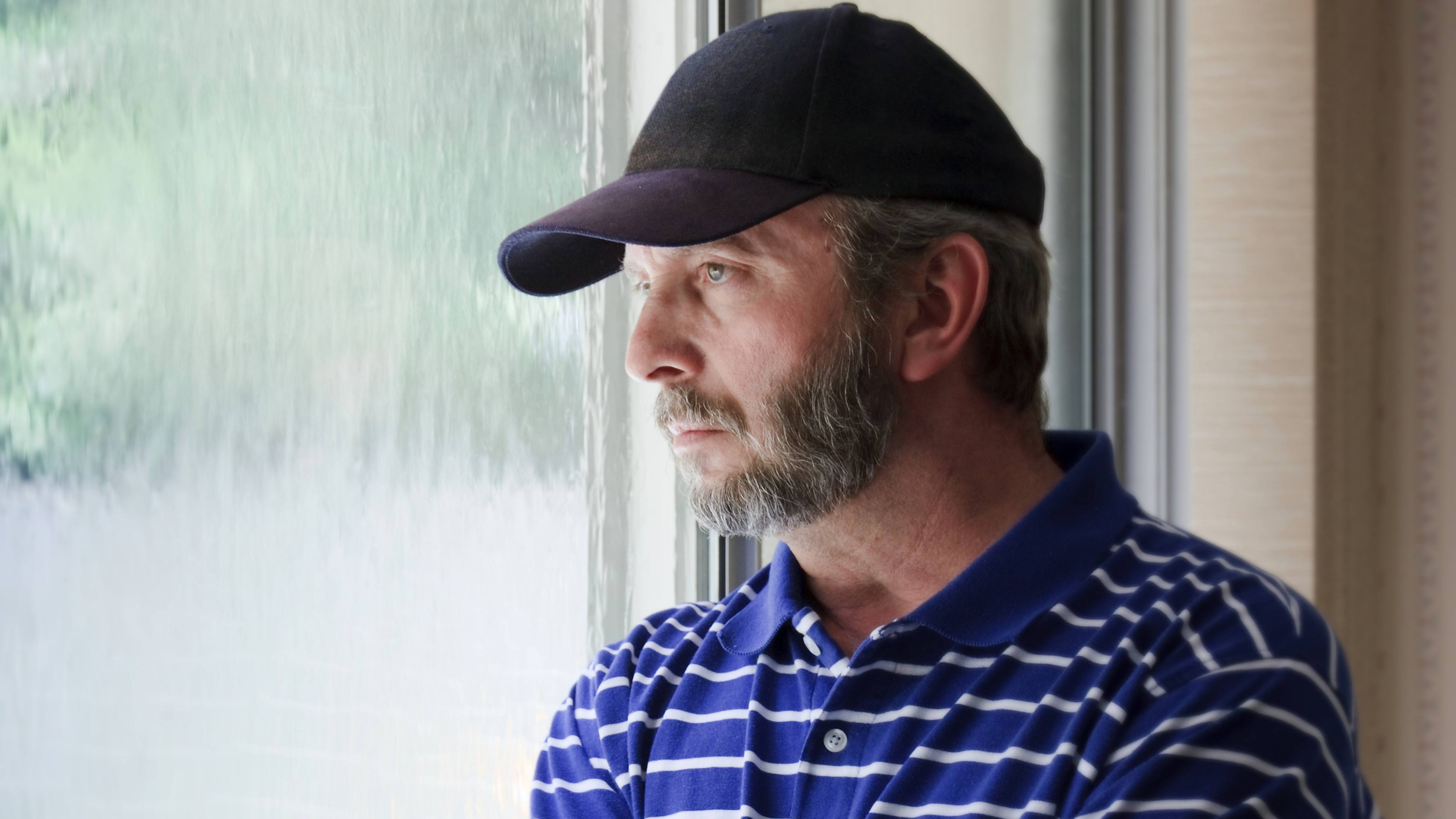 a middle-aged man wearing a baseball cap and staring out a window looking sad, thoughtful, depressed, worried