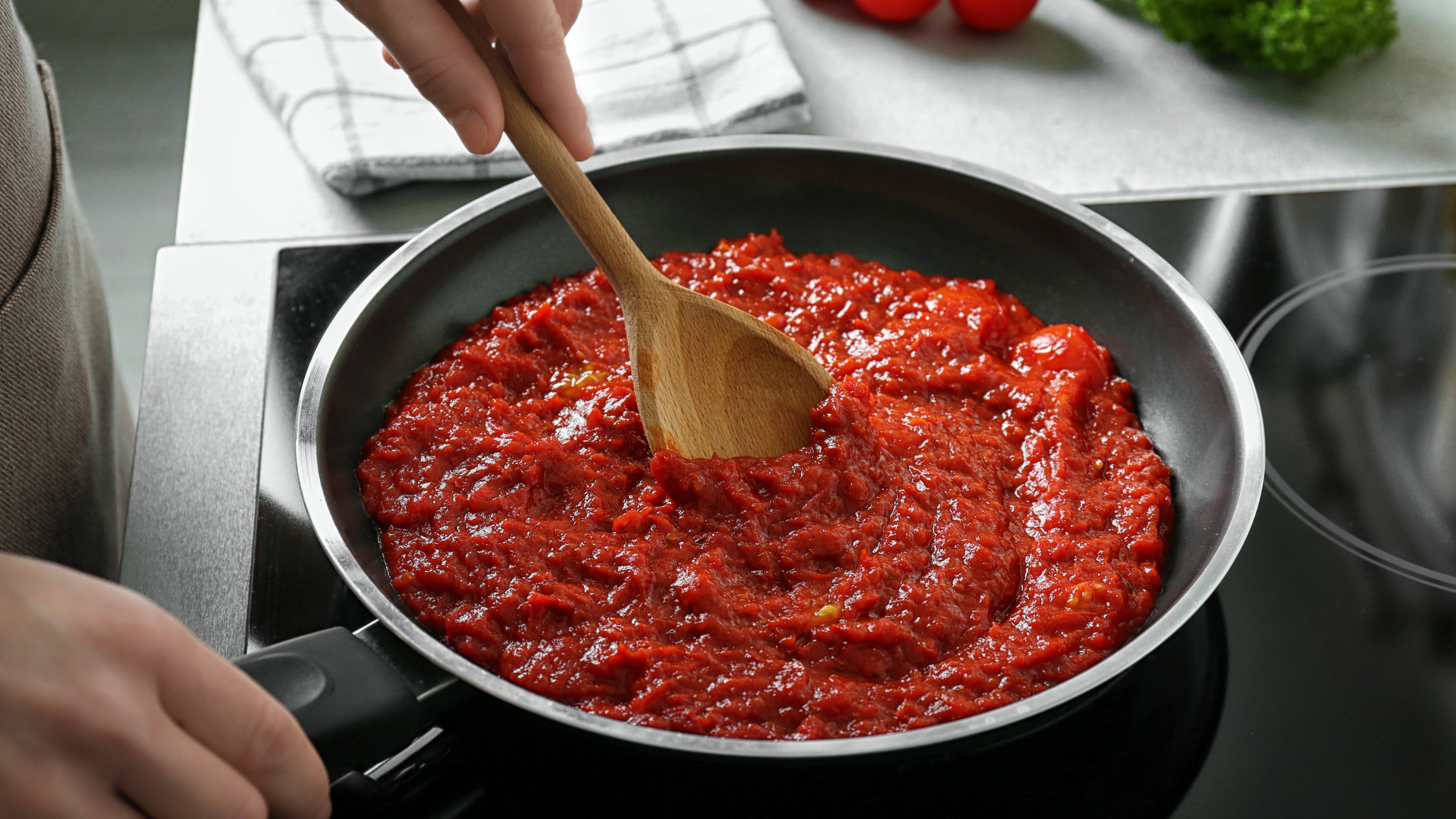 close up of a large frying pan with person at the stove cooking, stirring a red spaghetti or marinara sauce