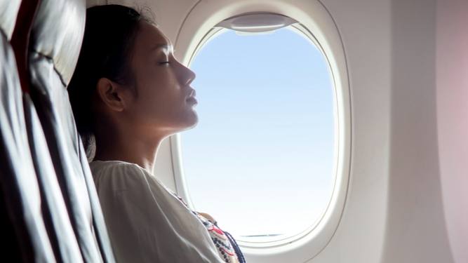 A young women sitting near a window on an airplane, with her eyes closed, resting or napping