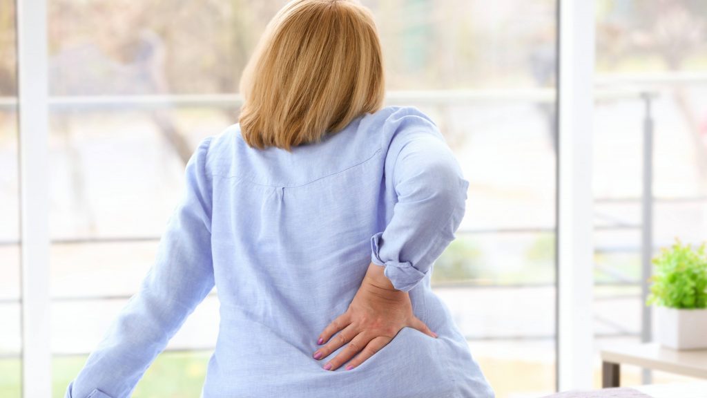 a middle-aged woman sitting on a bed with her hand on her back revealing pain