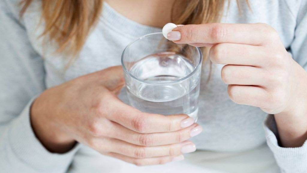 a young woman holding a glass of water and an aspirin or pain medicine tablet in her finger getting ready to drink and swallow it