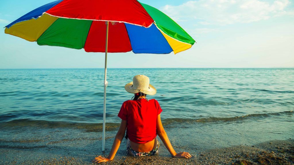 Una adolescente está sentada al sol en la playa, bajo una sombrilla y con un sombrero en la cabeza