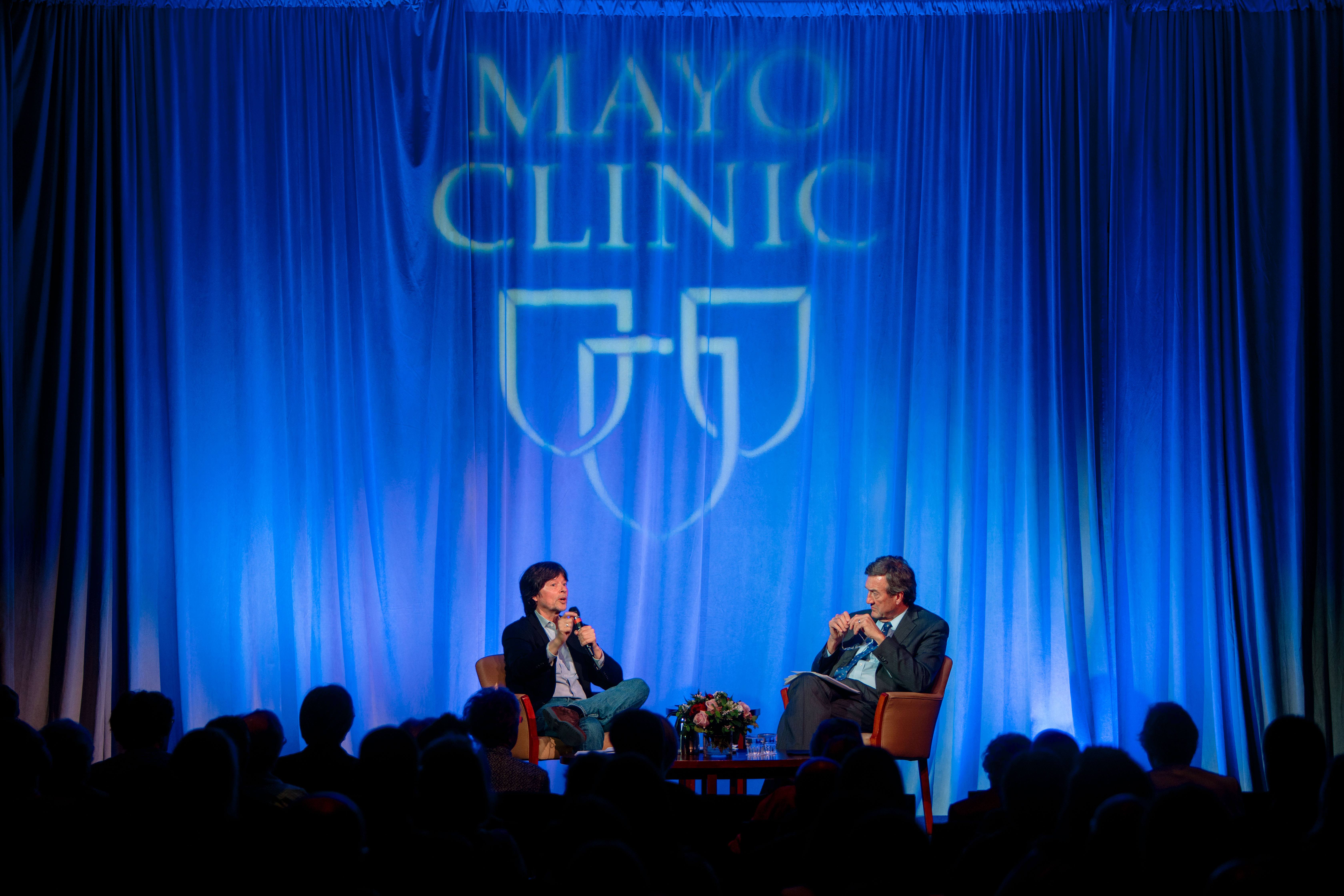 Photo of two males, Ken Burns and Dr. John Noseworthy on stage with a blue backdrop with the Mayo Clinic Logo of three shields