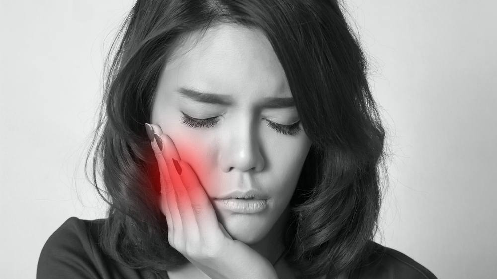 Una mujer joven presiona su mejilla con expresión de dolor porque tiene un dolor de muelas terrible.