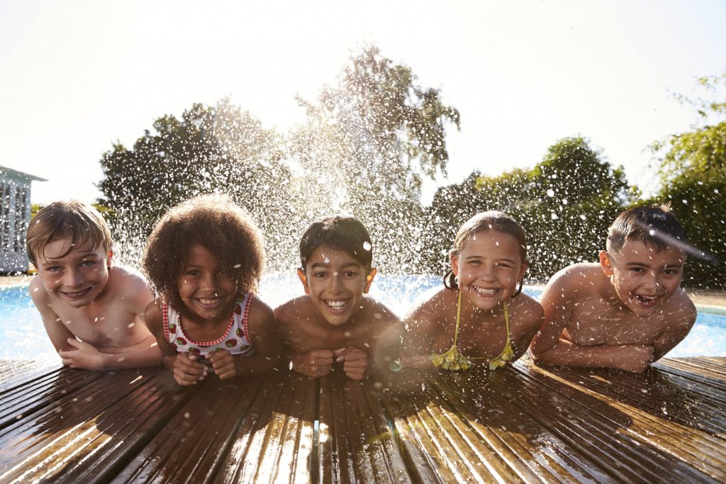 five smiling tweenage girls and boys leaning on the edge of a swimming pool with water splashing behind them