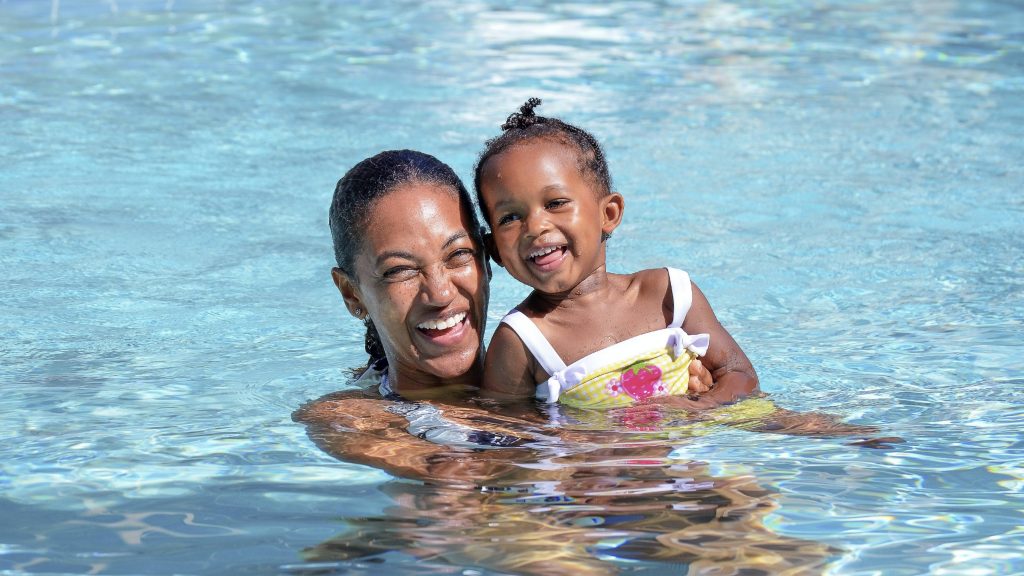 a little girl smiling as a woman, perhaps her mother, holds her in the water of a swimming pool