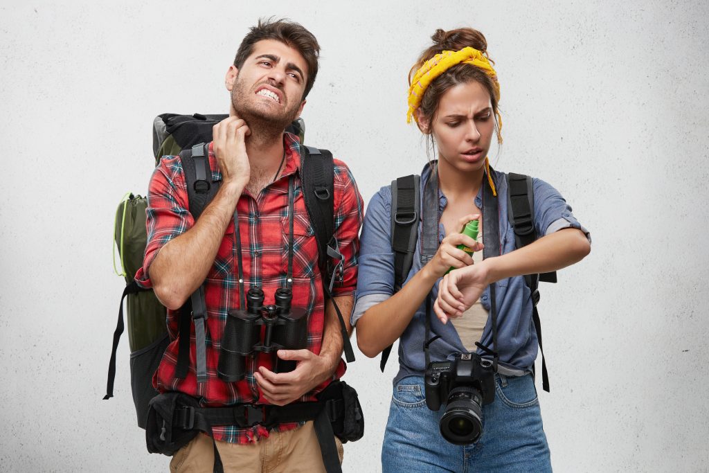a young couple dressed in hiking or camping clothes, looking distressed, scratching at insect bites